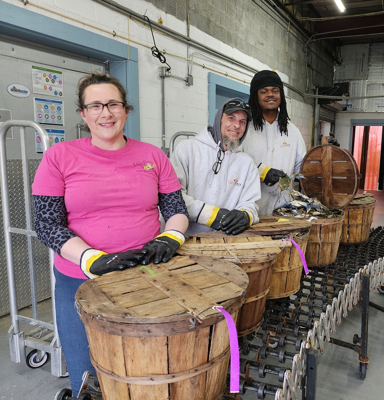 Three smiling Crab Place team members packing steamed crabs into wooden bushel baskets in the Crisfield warehouse