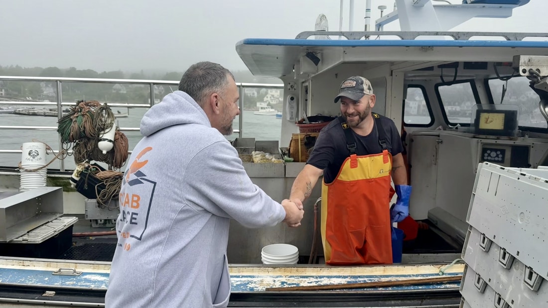 A Crab Place team member in a branded hoodie shaking hands with a Maine lobster boat captain in oil gear on the deck of the boat
