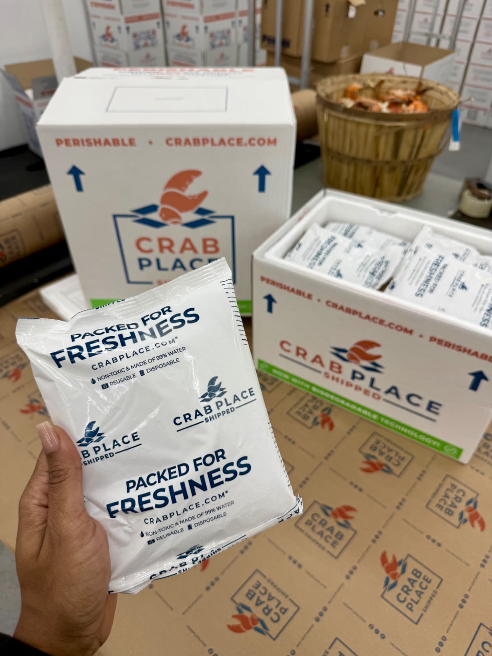 A hand holding a Crab Place Packed for Freshness cold pack in front of branded insulated shipping boxes in the warehouse.