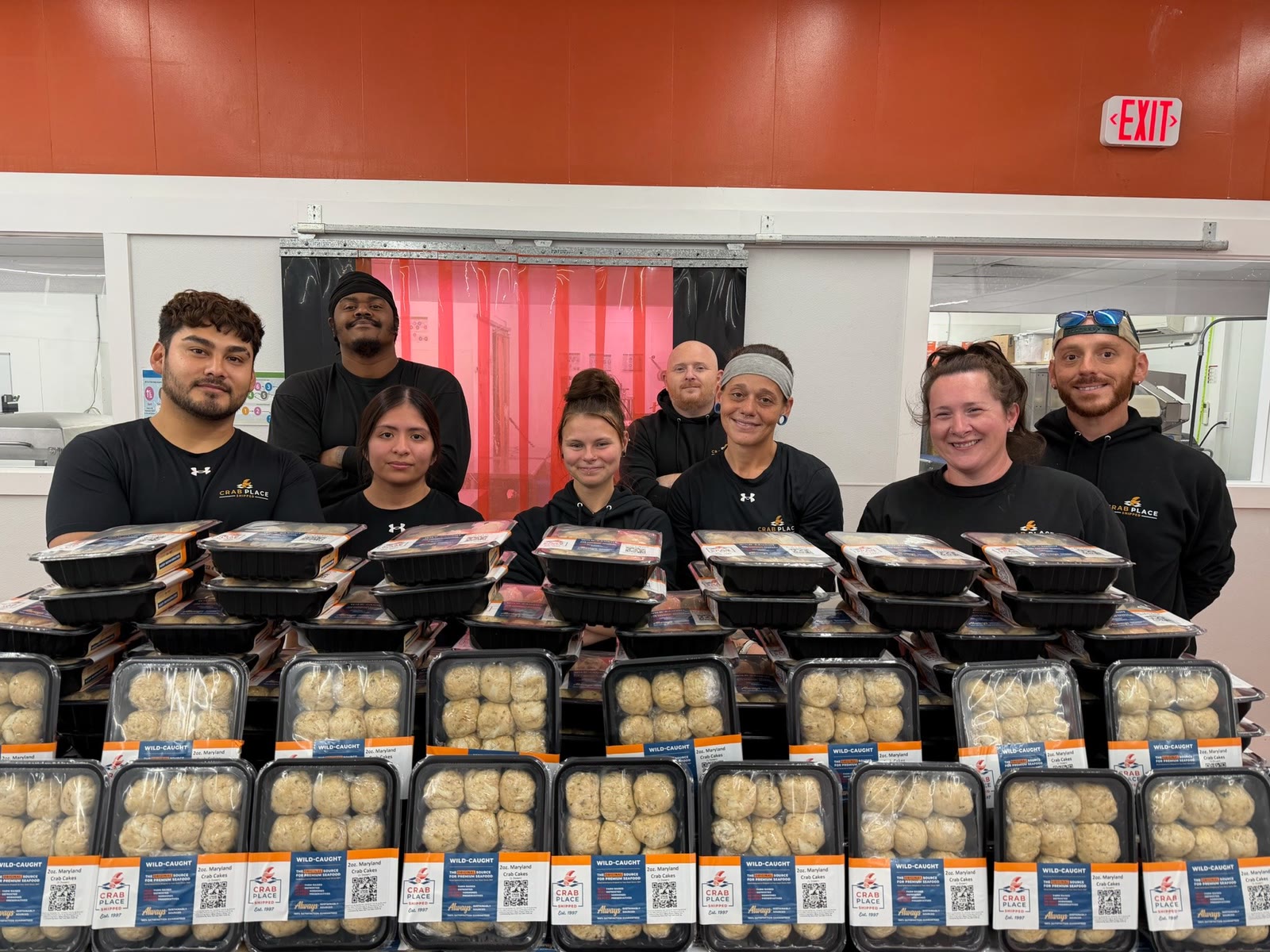 Eight Crab Place staff members in branded black shirts standing together behind stacked trays of packaged crab cakes and crab balls in the production kitchen.