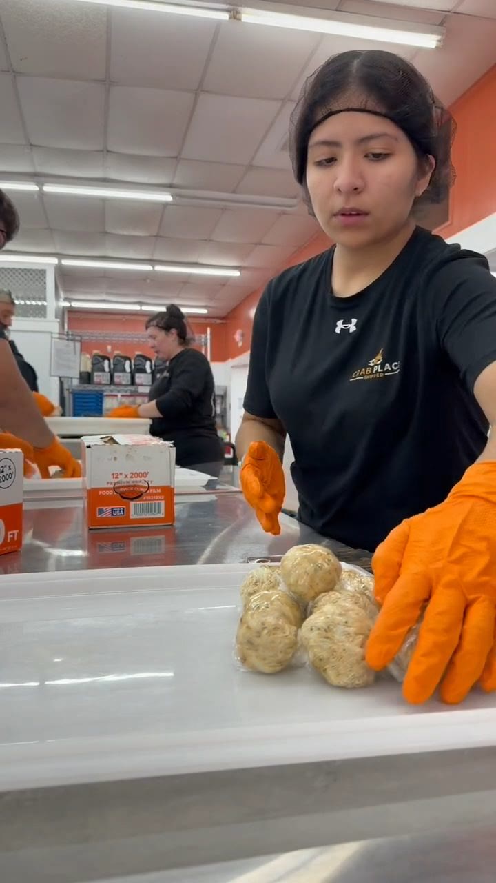 A Crab Place staff member in a branded shirt and orange gloves lining up freshly formed crab cakes on a stainless steel table.