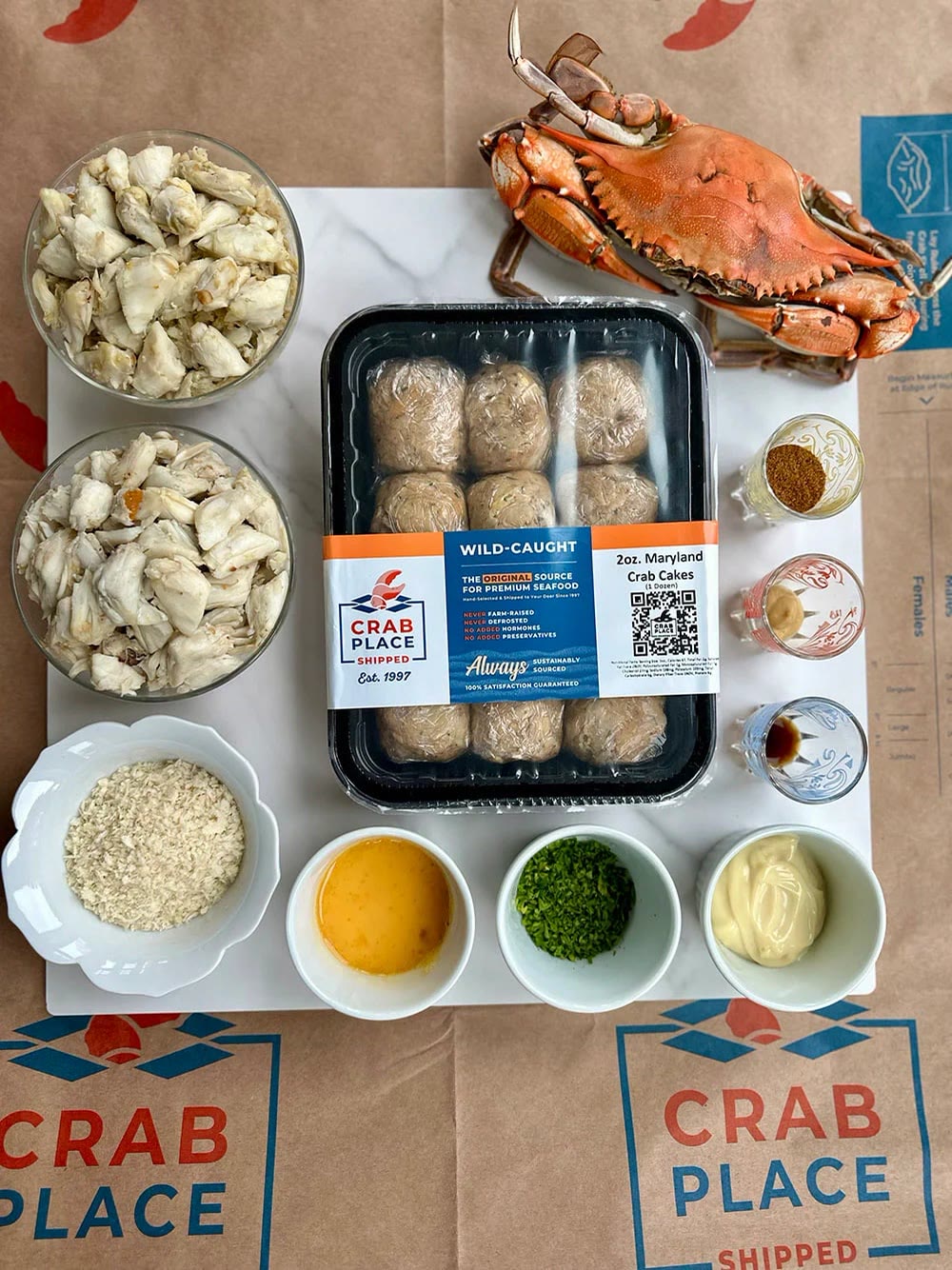 Overhead view of crab cake ingredients on Crab Place branded paper: bowls of jumbo lump and back fin crab meat, breadcrumbs, egg yolk, chopped parsley, mayonnaise, seasonings, a tray of 2 oz crab cakes and a steamed blue crab.