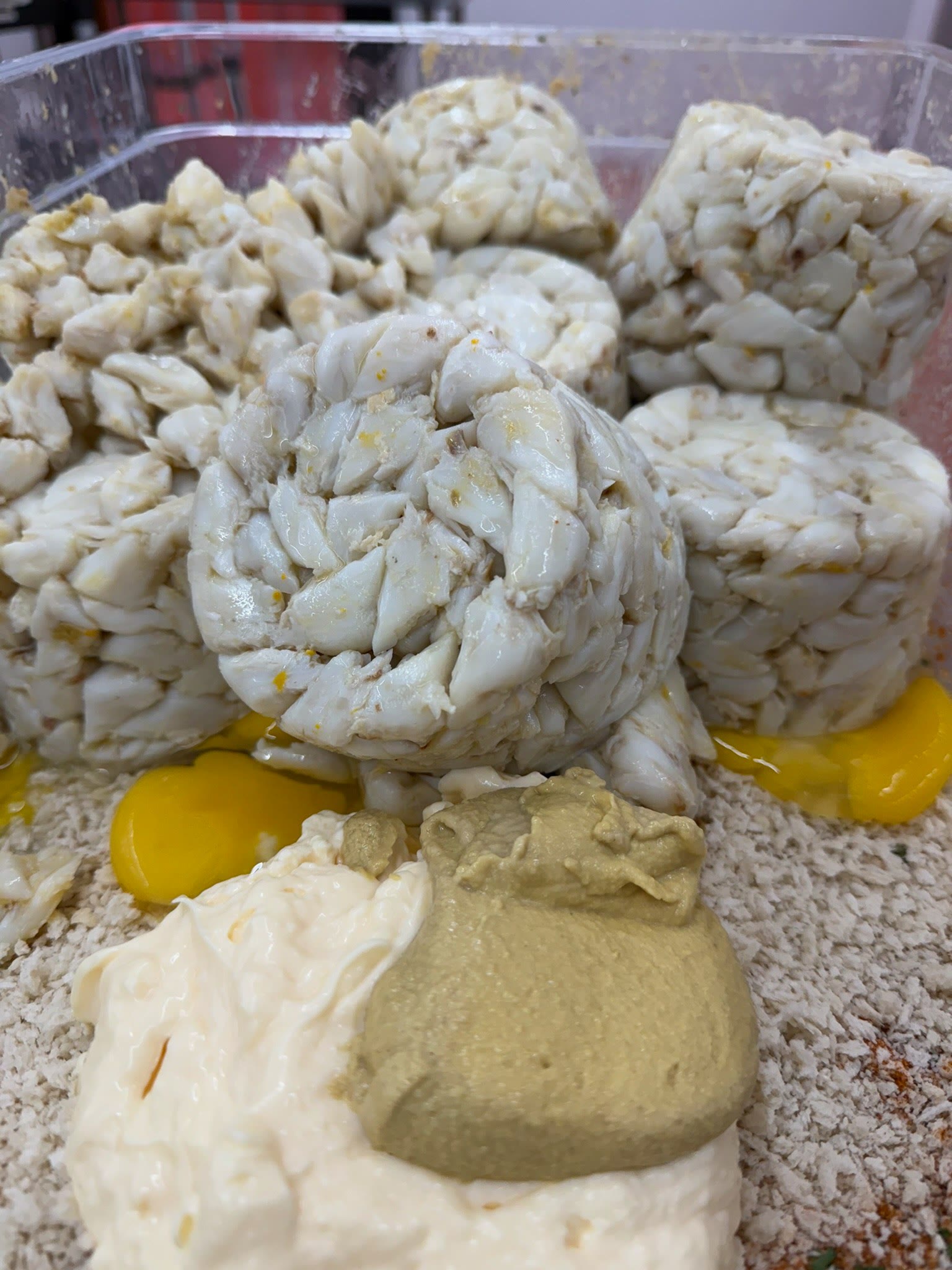 Close up of whole lumps of white crab meat sitting on top of egg yolk, mustard and breadcrumb binder in a clear tub before mixing.