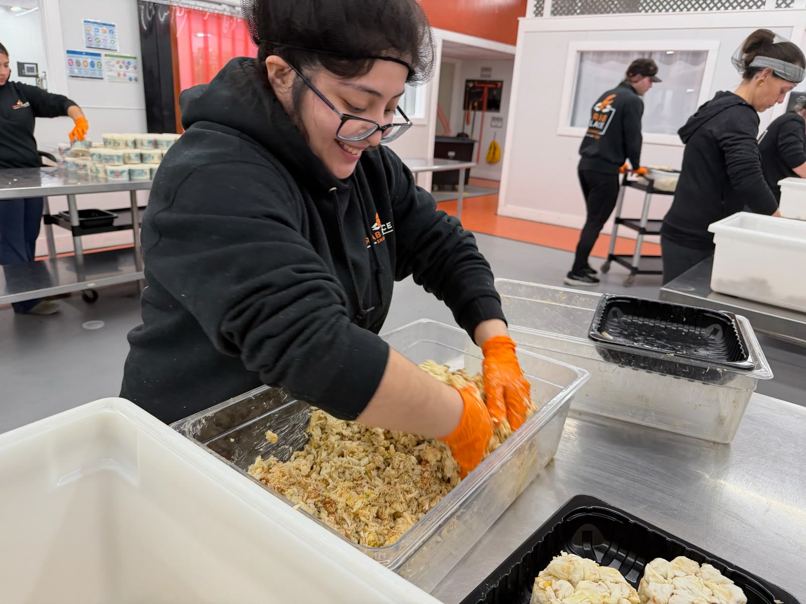 A Crab Place staff member in a branded hoodie and orange gloves smiling while mixing crab cake batter by hand in a clear tub in the production kitchen.