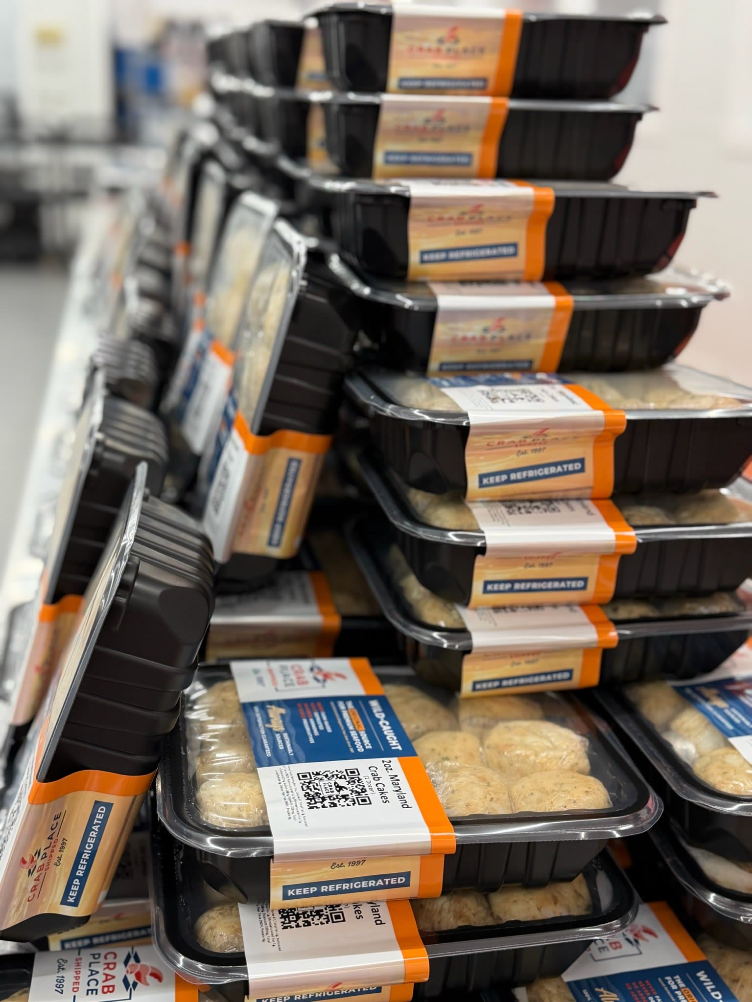 Stacked black trays of packaged Crab Place crab cakes with orange branded labels, ready for the freezer.