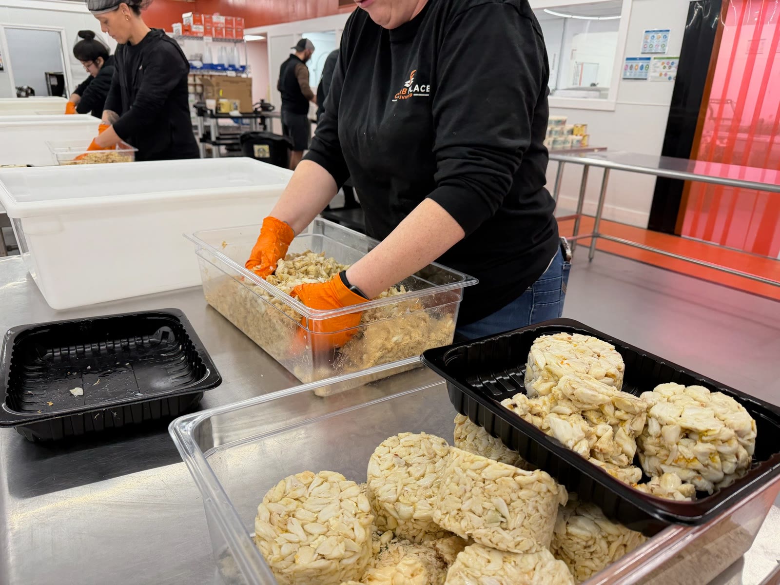 Crab Place staff in orange gloves portioning crab meat from clear tubs, with black trays of formed crab cakes in the foreground.