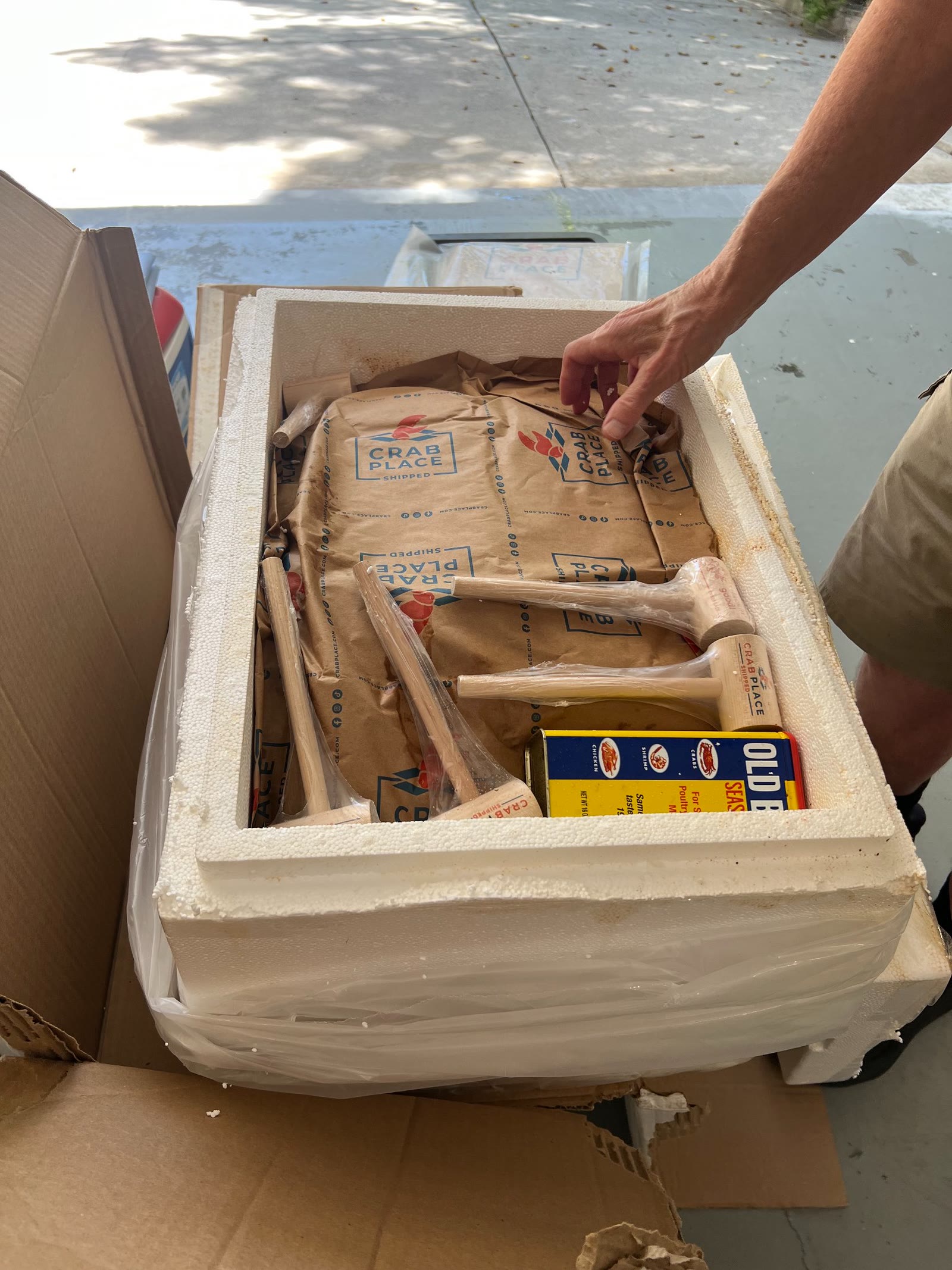 A customer opening an insulated Crab Place shipping box on a driveway, showing wrapped crab cakes, seasoning and a mallet packed inside.