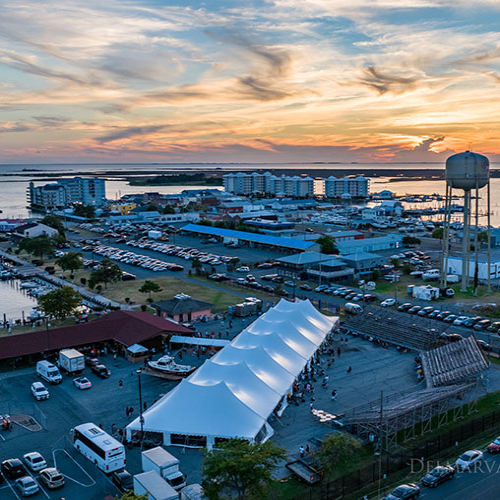 Aerial view at sunset of a large white event tent on the Crisfield waterfront, with the marina, the town and the red-crab water tower behind it under an orange sky.