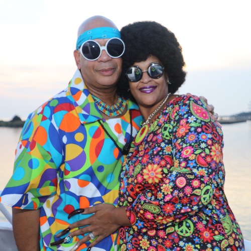 A couple in bright patterned outfits, round sunglasses and peace-sign prints, posing together by the water at sunset on Motown night at Crab and Cruise.