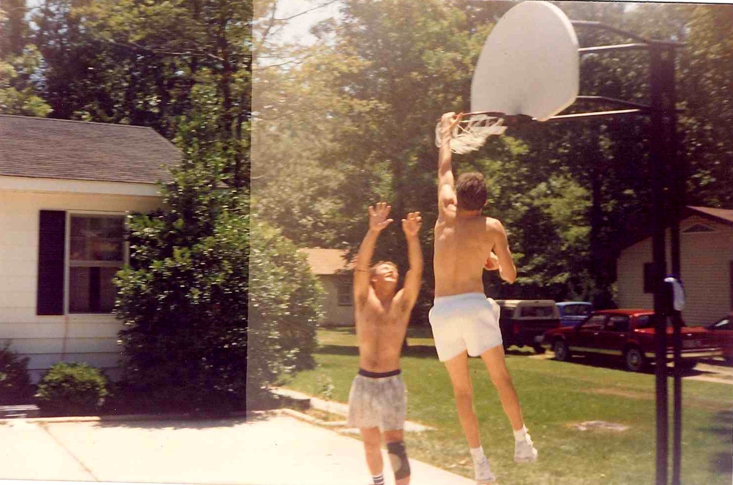 Two young men playing basketball at a backyard hoop on a summer day, one reaching for the rim.