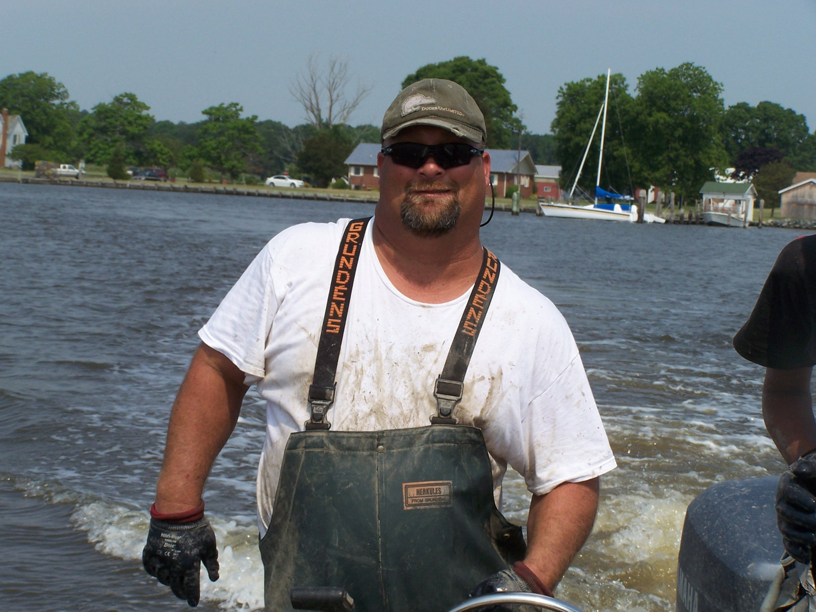 Waterman Moose Labo at the wheel of his boat in waders and a ball cap, with the Crisfield shoreline behind him.