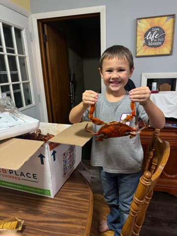 A young boy grinning and holding up a crab beside a Crab Place box