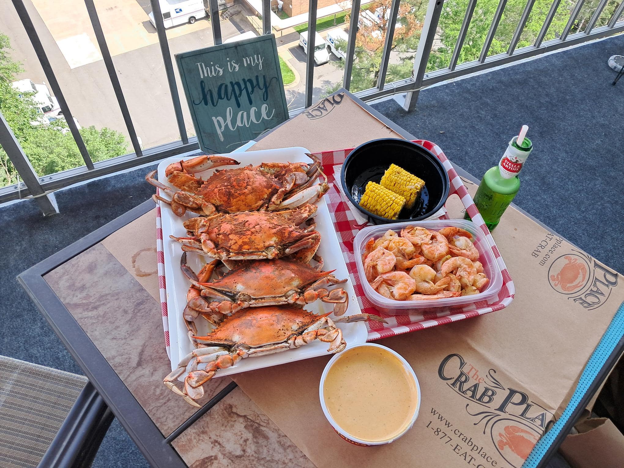 A balcony table set with Crab Place paper: a tray of steamed crabs, a tub of shrimp, corn on the cob and a sign that reads This is my happy place