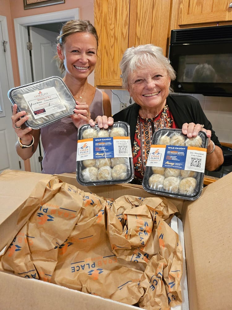 Two women in a kitchen holding up containers of Crab Place crab cakes